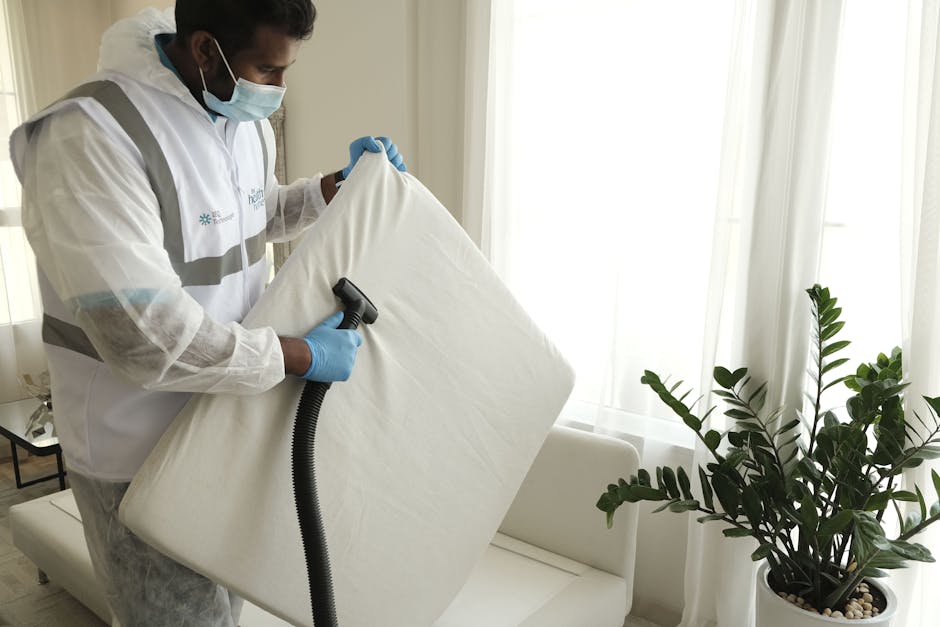 A professional cleaner from Lampton Carpet Cleaners is dressed in a white protective jumpsuit, blue gloves, and a face mask, using a handheld vacuum or upholstery cleaning device to perform deep clean